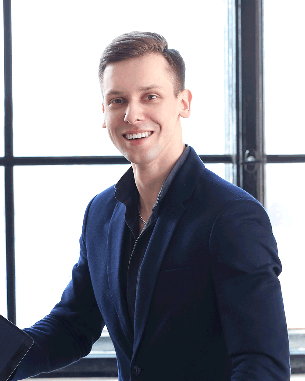 A smiling man holding a laptop in front of a window.