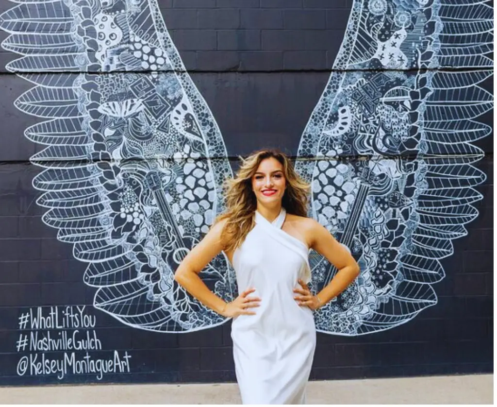 A woman in a white dress standing in front of a mural with wings.